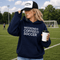 Woman wearing a navy hoodie with 'Weekends Coffee Soccer' text, holding a coffee cup on a soccer field.