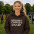 Woman wearing a hoodie with 'Weekends Coffee Soccer' text on a soccer field.