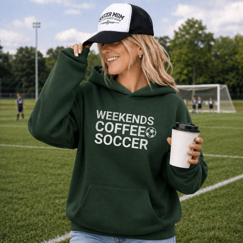 Woman on a soccer field wearing a hoodie and cap with text, holding a coffee cup.