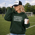 Woman on a soccer field wearing a hoodie and cap with text, holding a coffee cup.
