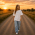 Woman wearing a white Comfort Colors 1717 t-shirt featuring a Prairie Roads Where the Wildflowers Grow prairie sunset wildflower back graphic on a gravel road at sunset.