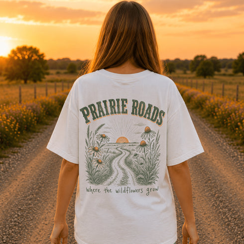 Woman wearing a white Comfort Colors 1717 t-shirt featuring a Prairie Roads Where the Wildflowers Grow prairie sunset wildflower back graphic on a gravel road at sunset.
