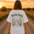 Woman wearing a white Comfort Colors 1717 t-shirt featuring a Prairie Roads Where the Wildflowers Grow prairie sunset wildflower back graphic on a gravel road at sunset.