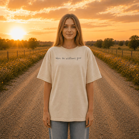 Woman wearing a white Comfort Colors 1717 t-shirt featuring a Prairie Roads Where the Wildflowers Grow prairie sunset wildflower back graphic on a gravel road at sunset.