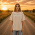 Woman wearing a white Comfort Colors 1717 t-shirt featuring a Prairie Roads Where the Wildflowers Grow prairie sunset wildflower back graphic on a gravel road at sunset.