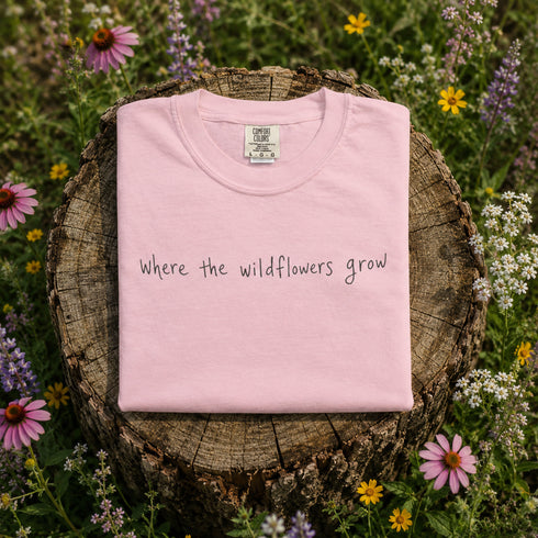 Pink t-shirt with 'Where the wildflowers grow' text on a wooden stump surrounded by wildflowers