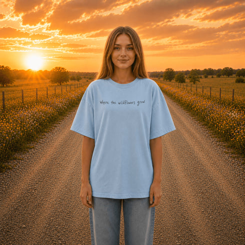 Woman wearing a white Comfort Colors 1717 t-shirt featuring a Prairie Roads Where the Wildflowers Grow prairie sunset wildflower back graphic on a gravel road at sunset.