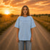Woman wearing a white Comfort Colors 1717 t-shirt featuring a Prairie Roads Where the Wildflowers Grow prairie sunset wildflower back graphic on a gravel road at sunset.