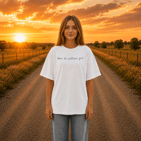 Woman wearing a white Comfort Colors 1717 t-shirt featuring a Prairie Roads Where the Wildflowers Grow prairie sunset wildflower back graphic on a gravel road at sunset.