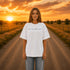 Woman wearing a white Comfort Colors 1717 t-shirt featuring a Prairie Roads Where the Wildflowers Grow prairie sunset wildflower back graphic on a gravel road at sunset.