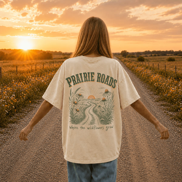 Woman wearing a white Comfort Colors 1717 t-shirt featuring a Prairie Roads Where the Wildflowers Grow prairie sunset wildflower back graphic on a gravel road at sunset.