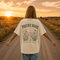 Woman wearing a white Comfort Colors 1717 t-shirt featuring a Prairie Roads Where the Wildflowers Grow prairie sunset wildflower back graphic on a gravel road at sunset.