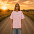 Woman wearing a white Comfort Colors 1717 t-shirt featuring a Prairie Roads Where the Wildflowers Grow prairie sunset wildflower back graphic on a gravel road at sunset.