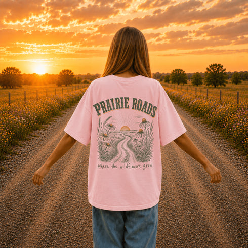 Woman wearing a white Comfort Colors 1717 t-shirt featuring a Prairie Roads Where the Wildflowers Grow prairie sunset wildflower back graphic on a gravel road at sunset.