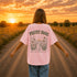 Woman wearing a white Comfort Colors 1717 t-shirt featuring a Prairie Roads Where the Wildflowers Grow prairie sunset wildflower back graphic on a gravel road at sunset.