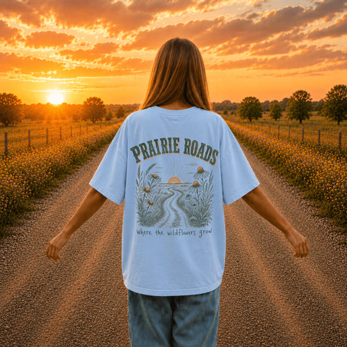 Woman wearing a white Comfort Colors 1717 t-shirt featuring a Prairie Roads Where the Wildflowers Grow prairie sunset wildflower back graphic on a gravel road at sunset.