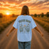 Woman wearing a white Comfort Colors 1717 t-shirt featuring a Prairie Roads Where the Wildflowers Grow prairie sunset wildflower back graphic on a gravel road at sunset.