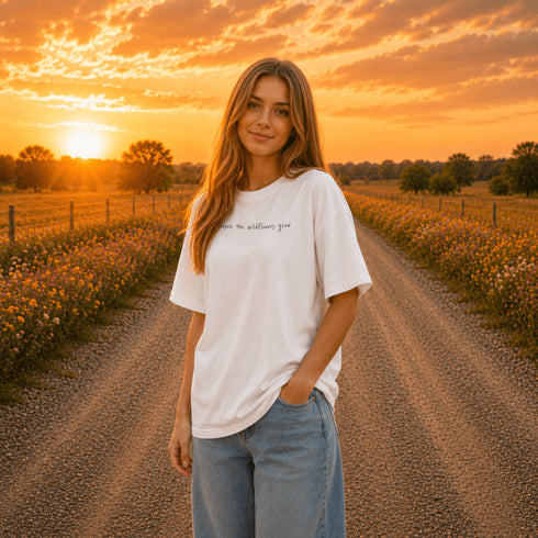 Woman wearing a white Comfort Colors 1717 t-shirt featuring a Prairie Roads Where the Wildflowers Grow prairie sunset wildflower back graphic on a gravel road at sunset.