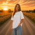 Woman wearing a white Comfort Colors 1717 t-shirt featuring a Prairie Roads Where the Wildflowers Grow prairie sunset wildflower back graphic on a gravel road at sunset.