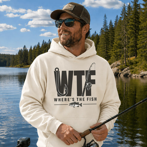 A man wearing a cream-colored hoodie with a black graphic that reads 'WTF WHERE'S THE FISH' stands on a lakeshore with trees and water in the background.