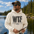 A man wearing a cream-colored hoodie with a black graphic that reads 'WTF WHERE'S THE FISH' stands on a lakeshore with trees and water in the background.