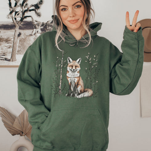 This hoodie features a stunning graphic of a vibrant red fox poised amidst the shadows and light of a lush woodland scene.