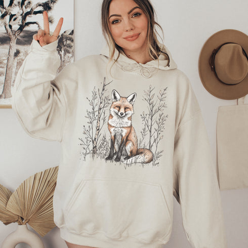 This hoodie features a stunning graphic of a vibrant red fox poised amidst the shadows and light of a lush woodland scene.