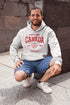 Woman wearing grey Canada Always Strong hoodie with red maple leaf graphic and “Est. 1867” design, styled outdoors in a natural setting