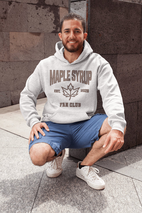 Man wearing a Maple Syrup Fan Club hoodie with maple leaf graphic, smiling outdoors in a natural setting with trees in the background